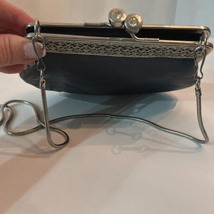 Pu&ograve; includere: Una pochette blu scuro con una montatura in metallo argentato e una tracolla a catena. La chiusura presenta due accenti decorativi rotondi e trasparenti. La borsa ha un aspetto strutturato e un design decorativo in filigrana argentata.