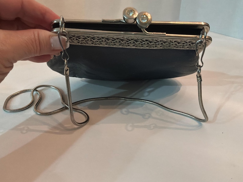 Pu&ograve; includere: Una pochette blu scuro con una montatura in metallo argentato e una tracolla a catena. La chiusura presenta due accenti decorativi rotondi e trasparenti. La borsa ha un aspetto strutturato e un design decorativo in filigrana argentata.