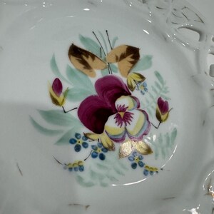 May include: White porcelain dish with a floral design. The center features a purple and yellow pansy with green leaves, brown accents, and small blue flowers. The rim has a delicate, openwork design.