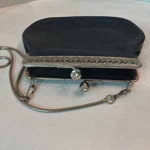 Pu&ograve; includere: Una pochette vintage in raso nero con montatura e catena in metallo argentato. La montatura presenta un design traforato ornato. La pochette ha una chiusura a scatto e una piccola catena argentata.