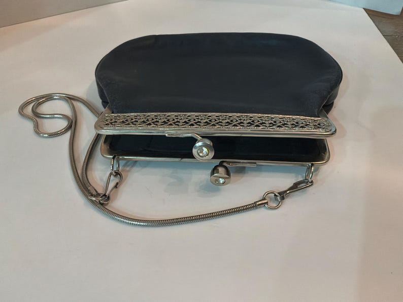 Pu&ograve; includere: Una pochette vintage in raso nero con montatura e catena in metallo argentato. La montatura presenta un design traforato ornato. La pochette ha una chiusura a scatto e una piccola catena argentata.