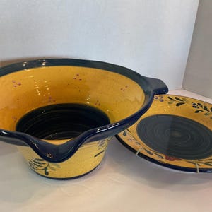 May include: A ceramic bowl with a wide, flared rim and a matching plate. The bowl features a deep blue interior base and rim, with a yellow body decorated with small purple speckles and green leaf accents. The plate has a matching blue center.
