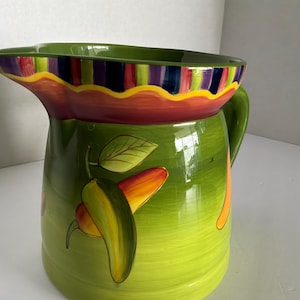 May include: A ceramic pitcher with a glossy lime green finish, featuring a painted design of yellow and orange peppers with green leaves. The rim is decorated with a wide band of vertical purple, blue, and red stripes, separated by a wavy yellow line.