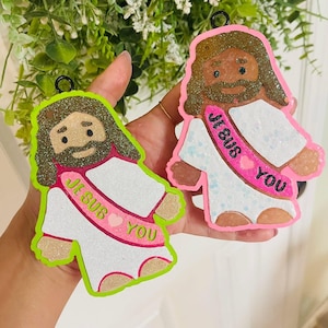 May include: Two glittery Jesus ornaments. One has a neon green border, the other a pink border. Both have brown hair and beards, white robes, and pink sashes that read "JESUS YOU".