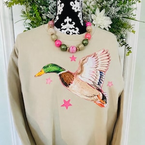 May include: A beige crewneck sweatshirt featuring a hand-painted mallard duck in flight with a green head, brown chest, and white-tipped wings. The design includes several bright pink stars surrounding the duck. A beaded necklace with pink, gold, and olive green spheres is worn around the neck of the display form.