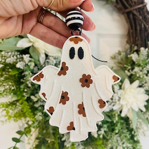 May include: A white glittery ghost ornament featuring brown flower patterns and black oval eyes. The ghost is suspended by a black string attached to a round bead with black and white horizontal stripes, designed for hanging as a decorative accent.