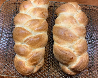May include: Two golden brown braided loaves of bread on a wire rack.