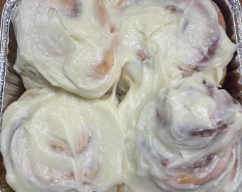 May include: Four golden-brown cinnamon rolls arranged in a square aluminum foil pan. Each roll is topped with a thick layer of white cream cheese frosting that swirls into the crevices of the dough, creating a cohesive, glazed pastry surface.