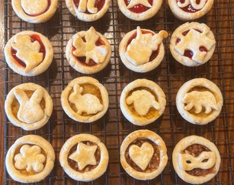 May include: A cooling rack with 16 mini pies. The pies have various designs, including a heart, a star, a shamrock, a bunny, a pie, a number 36, a star, a circle, a maple leaf, a puzzle piece, a state of Missouri, and a mask.
