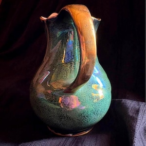 Può includere: Una brocca verde e oro con un grande manico dorato curvo. La brocca ha una consistenza maculata e una lucentezza iridescente, che riflette vari colori. Anche la base e il bordo sono rifiniti in oro.