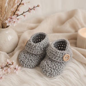 Könnte beinhalten: Ein Paar handgefertigte graue Häkel-Babyschuhe mit strukturiertem Stichmuster und einem einzelnen hellen Holzknopf an der Seite jedes Schuhs. Die Schuhe haben ein umgeschlagenes Bündchen und eine abgerundete Zehenpartie aus weichem Garn.