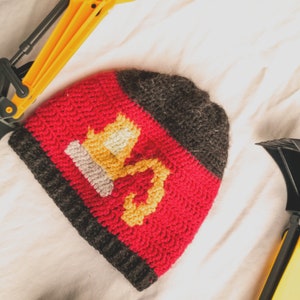 May include: A red, grey and yellow knitted hat with a crown design. The hat is lying on a white surface next to two toy construction vehicles.