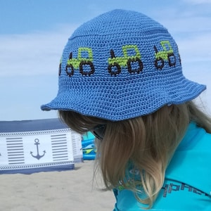 Könnte beinhalten: Ein blauer Häkelhut mit breiter Krempe und einem Muster aus grünen Traktoren auf der Vorderseite.