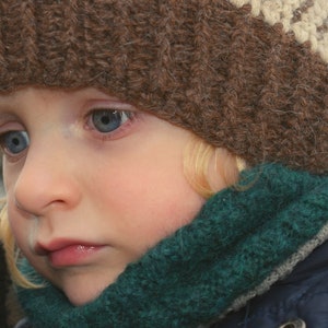 Könnte beinhalten: Ein kleines Kind trägt eine braune Strickmütze mit weißem Rand und einen grünen Strick Schal. Das Kind hat blonde Haare und blaue Augen.