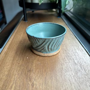 May include: A teal ceramic bowl with a textured, wavy design. The bowl is sitting on a wooden surface.