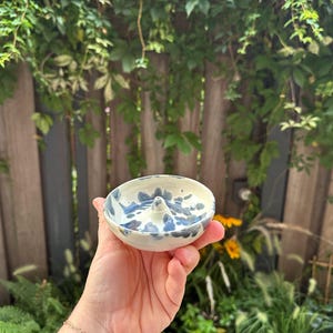 Puede incluir: Un pequeño plato de cerámica blanco con diseños abstractos azules. El plato tiene un centro elevado para sujetar anillos y se sostiene en una mano sobre un fondo de vegetación y una valla de madera.