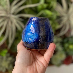May include: A small, hand-thrown ceramic vase with a blue and black glaze. The vase has a rounded, bulbous shape and a slightly uneven rim.