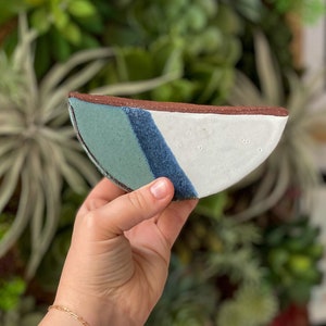 Puede incluir: Un plato de cerámica en forma de semicírculo con un esmalte blanco, azul y verde. El plato tiene un borde ligeramente redondeado y un acabado liso y brillante.