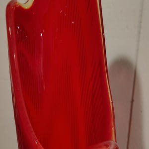 May include: A tall, glossy red vase with a distinctive, curved form. The vase features a textured surface with vertical lines and a cream-coloured rim. The design is modern and striking, suitable for home decor.