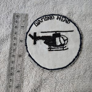 May include: A circular patch with a white background and a black helicopter design. The text "DIAMOND HEAD" is embroidered in black around the top edge. A ruler is placed next to the patch.
