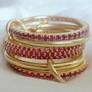 Può includere: Una pila di anelli color oro, alcuni adornati con piccole e vibranti pietre preziose rosse. Gli anelli sono disposti in un design a strati, creando un aspetto visivamente accattivante ed elegante. Lo sfondo è un morbido tessuto bianco.