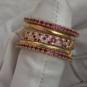 Op de afbeelding: Een verzameling gouden ringen, sommige versierd met kleine, levendige roze edelstenen, andere met heldere, sprankelende stenen. De ringen zijn op een vinger gestapeld en tonen hun verschillende ontwerpen en texturen. De algehele esthetiek is er een van luxe en elegantie.