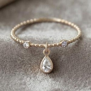 May include: A gold-toned ring featuring a delicate beaded band with two small round clear stones set on either side. A single pear-shaped clear stone hangs from the center of the band, creating a dangling drop effect on this fine jewelry piece.