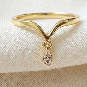 Puede incluir: Un anillo de oro en forma de V con una piedra preciosa transparente de talla marquesa que cuelga del punto central. La banda tiene un acabado liso y pulido, y la pequeña piedra está engastada en un delicado marco de oro, creando una pieza sencilla.