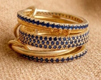 Può includere: Una pila di tre anelli dorati con pietre blu incastonate. Gli anelli superiore e inferiore sono fasce sottili con una fila di pietre blu, mentre l'anello centrale è una fascia più larga con diverse file di piccole pietre blu rotonde.