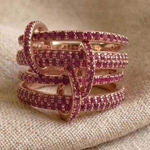 May include: A rose gold ring featuring multiple bands connected by a vertical bar, all encrusted with small, vibrant pink stones. The design creates a layered, interconnected appearance with a polished metallic finish and uniform gemstone setting.