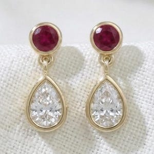 May include: A pair of gold drop earrings featuring a round, deep red ruby gemstone at the top, connected to a larger, pear-shaped clear white diamond-like stone below. Each stone is set in a smooth gold bezel setting with a delicate gold link.