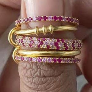 Può includere: Una collezione di anelli color oro, alcuni adornati con piccole pietre preziose rosa. Gli anelli sono impilati su un dito, mostrando il loro design. Le pietre preziose sembrano essere un mix di pietre rosa e chiare.