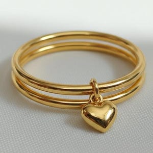 May include: A gold-colored ring with three bands and a small heart charm. The ring is made of a shiny, reflective metal and sits on a white, textured surface. The heart charm hangs from the bottom band.