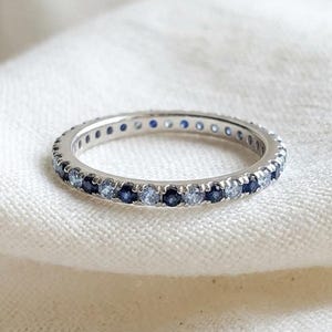 Puede incluir: Un anillo de banda fina en metal plateado que presenta una hilera continua de pequeñas piedras preciosas de corte redondo que alternan entre el azul marino profundo y el azul hielo pálido, creando un sutil efecto degradado en el anillo.