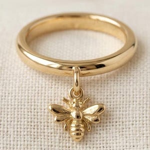 May include: A gold ring featuring a bee pendant. The ring has a smooth, rounded band, and the bee charm is intricately detailed. The ring is displayed on a textured, cream-colored surface.