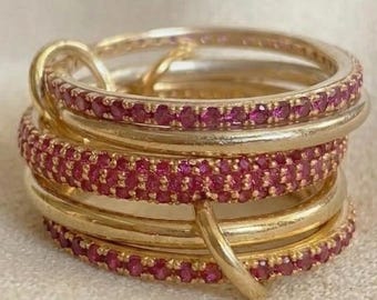 Può includere: Una pila di anelli color oro caratterizzata dall'alternanza di fasce in metallo liscio e lucido e fasce incastonate con piccole pietre rosa brillante. Gli anelli sono interconnessi, creando un gioiello circolare a strati e strutturato.