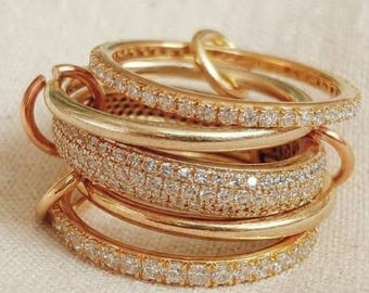 Può includere: Una pila di cinque anelli color oro. Tre anelli presentano un'incastonatura a pavé con piccole pietre chiare che circondano la fascia, mentre due anelli sono fasce lisce color oro. Gli anelli sono sovrapposti in un unico gruppo verticale.