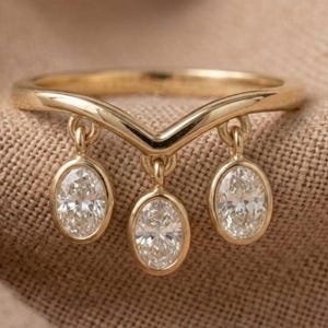 May include: A gold V-shaped ring featuring three dangling oval-cut clear stones. The band has a polished finish, and the three stones are suspended from the center and sides of the chevron-shaped band, creating a delicate and symmetrical appearance.