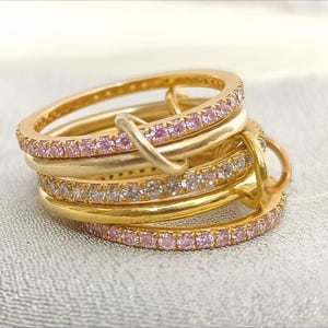 Op de afbeelding: Een stapel goudkleurige ringen met roze en heldere edelstenen. De ringen zijn in een gelaagd ontwerp gerangschikt, waarbij sommige ringen een doorlopende rij roze edelstenen hebben en andere heldere edelstenen. De ringen liggen op een lichtgekleurd oppervlak.