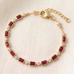 May include: A gold chain bracelet featuring alternating rectangular red baguette-cut stones and small round clear stones. The bracelet includes a lobster claw clasp and an adjustable extender chain with circular links at the end of the piece.