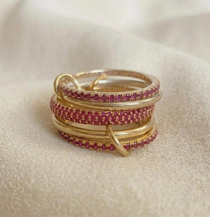 Pu&ograve; includere: Una pila di anelli color oro caratterizzata dall'alternanza di fasce in metallo liscio e lucido e fasce incastonate con piccole pietre rosa brillante. Gli anelli sono interconnessi, creando un gioiello circolare a strati e strutturato.