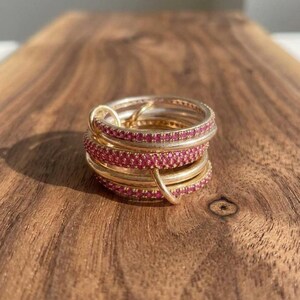 Pu&ograve; includere: Una pila di anelli color oro composta da diverse fasce, alcune delle quali incastonate con piccole pietre rosa brillante. Gli anelli sono collegati tra loro da un piccolo occhiello color oro, creando un gioiello circolare a strati.