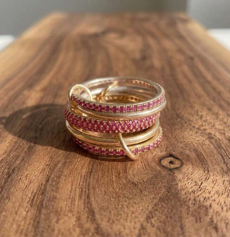 Pu&ograve; includere: Una pila di anelli color oro composta da diverse fasce, alcune delle quali incastonate con piccole pietre rosa brillante. Gli anelli sono collegati tra loro da un piccolo occhiello color oro, creando un gioiello circolare a strati.