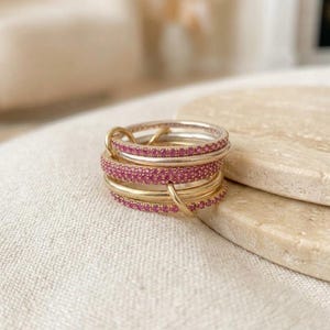 Pu&ograve; includere: Una pila di anelli intrecciati caratterizzata da fasce alternate in oro lucido e fasce incastonate con piccole pietre preziose rosa brillante. Gli anelli sono collegati da un piccolo anello d'oro, creando un gioiello circolare a pi&ugrave; strati.