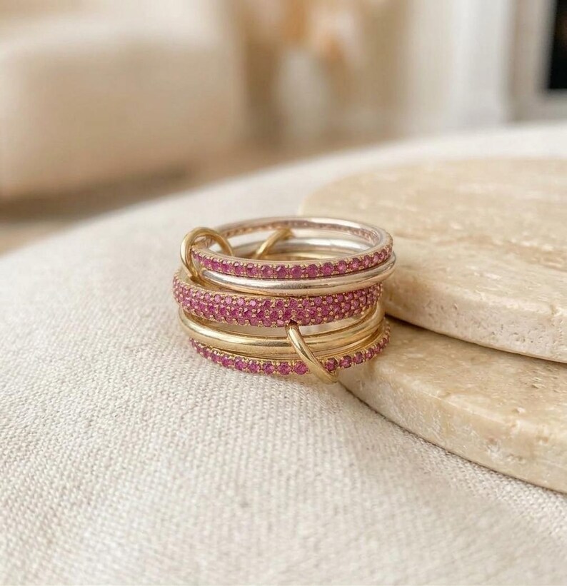 Pu&ograve; includere: Una pila di anelli intrecciati caratterizzata da fasce alternate in oro lucido e fasce incastonate con piccole pietre preziose rosa brillante. Gli anelli sono collegati da un piccolo anello d'oro, creando un gioiello circolare a pi&ugrave; strati.