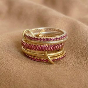 Pu&ograve; includere: Una pila di anelli dorati con fasce alternate di metallo liscio e fasce incastonate con piccole pietre rosa brillante. Gli anelli sono collegati tra loro da un piccolo anello dorato, creando una composizione di gioielli a strati.