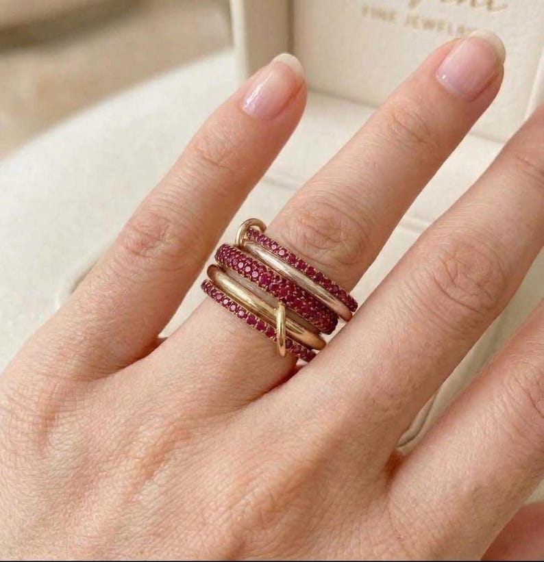 Pu&ograve; includere: Un anello a pi&ugrave; strati caratterizzato da fasce alternate in oro lucido e fasce incastonate con piccole pietre rosse vivaci. Il design &egrave; composto da diversi anelli interconnessi che si avvolgono attorno al dito, creando un aspetto sovrapposto con un mix di superfici metalliche lisce e segmenti testurizzati ricoperti di pietre preziose.