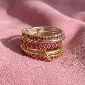 Pu&ograve; includere: Una pila di anelli color oro con pietre rosa incastonate. Gli anelli sono disposti in un gruppo verticale, con alcune fasce che presentano una fila continua di piccole gemme rosa circolari e altre con una finitura oro liscia e lucida.