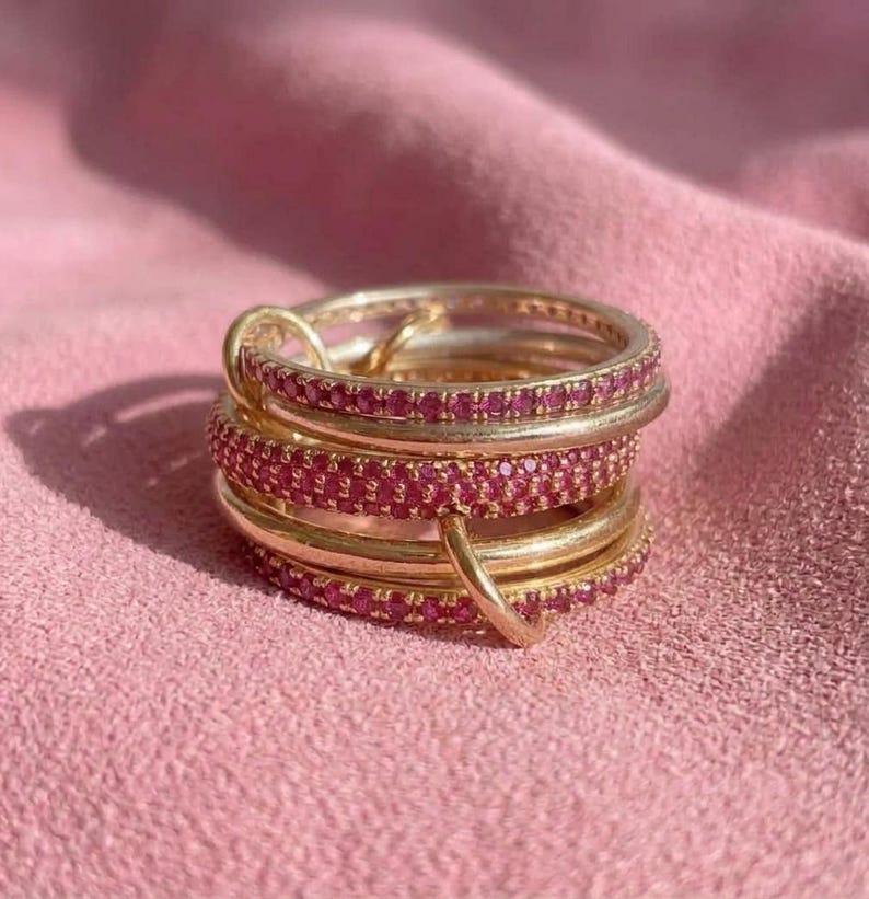 Pu&ograve; includere: Una pila di anelli color oro con pietre rosa incastonate. Gli anelli sono disposti in un gruppo verticale, con alcune fasce che presentano una fila continua di piccole gemme rosa circolari e altre con una finitura oro liscia e lucida.