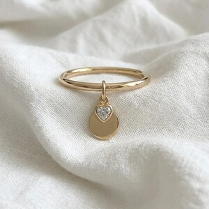 Könnte beinhalten: Ein goldener Ring mit einer dünnen, glatten Ringschiene und einem hängenden Anhänger. Der Anhänger besteht aus einem kleinen, klaren, herzförmigen Edelstein, der über einer flachen, runden goldenen Scheibe angebracht ist. Das Metall ist poliert.